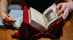 A person writing in a notepad with a pencil, next to an open book on a red fabric cushion.